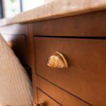 A close-up of a wooden cabinet with gold cup-style drawer pulls and a marble countertop. The warm wood tones contrast with the smooth, polished brass hardware, creating an elegant and classic aesthetic.