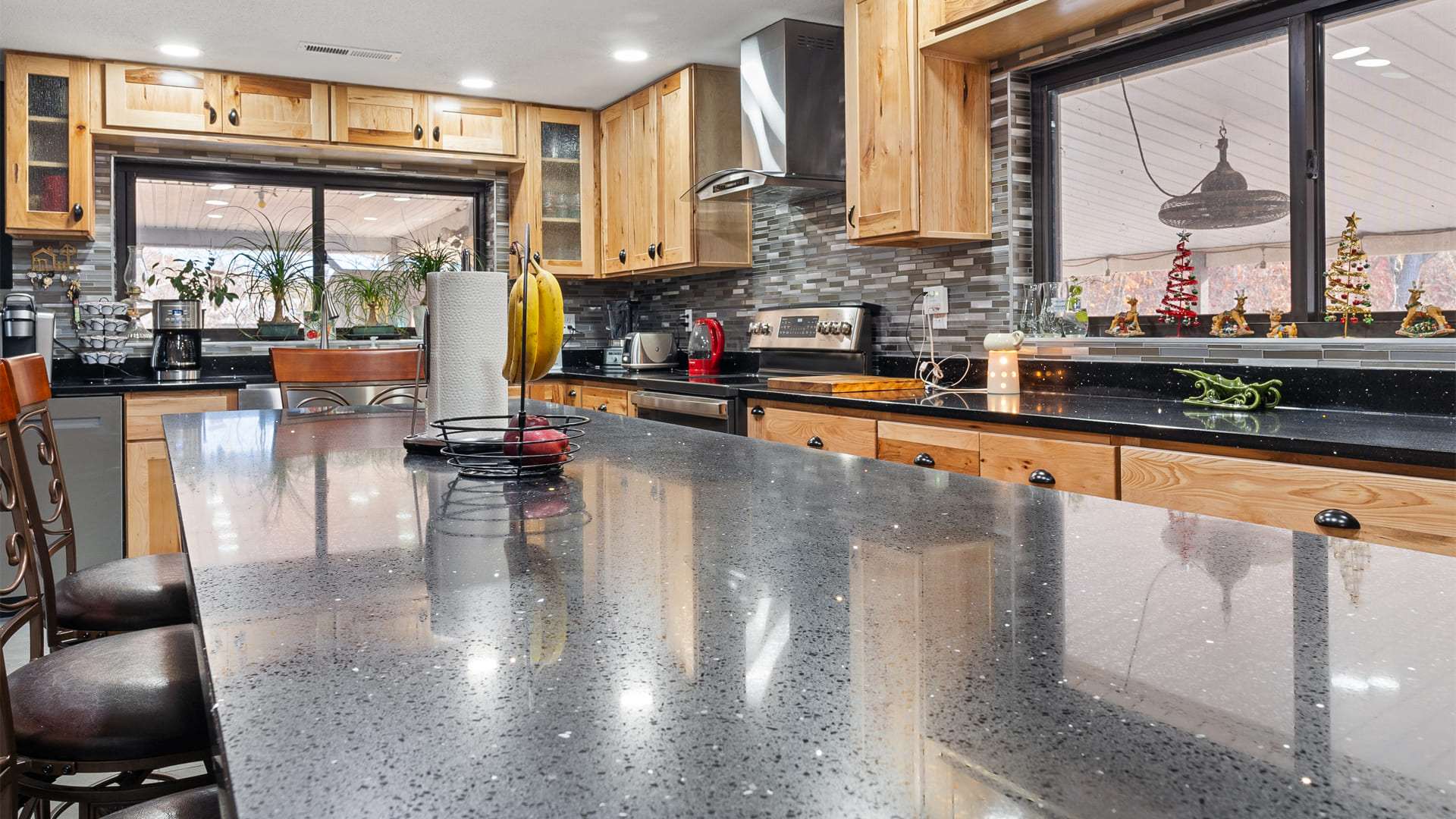 A spacious kitchen with a large countertop island at the center and rows of blonde wood cabinets set off with a stone, staggered tile backsplash
