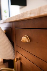 A close-up of a wooden cabinet with gold cup-style drawer pulls and a marble countertop. The warm wood tones contrast with the smooth, polished brass hardware, creating an elegant and classic aesthetic.