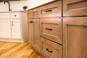 A close-up of light wood kitchen cabinets with black hardware, set against a contrasting background of white cabinetry with black handles. The natural wood grain and matte black pulls create a modern rustic aesthetic.